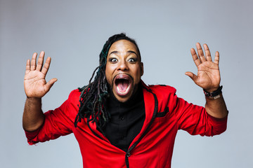 Studio portrait of a man wearing a red jacket suit and dreadlocks with both hands up and screaming