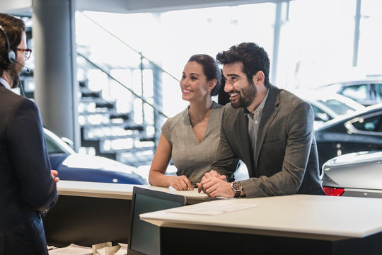 Couple customers talking to receptionist at desk in car dealership showroom