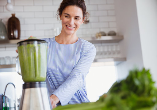 Smiling woman making healthy green smoothie in blender in kitchen