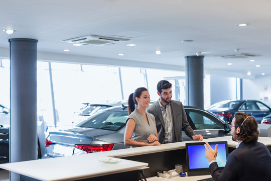 Couple customers talking to male receptionist at desk in car dealership showroom