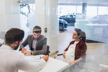 Car salesman explaining financial contract paperwork to pregnant couple customers in car dealership office