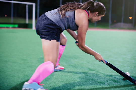 Determined young female field hockey player hitting the ball, playing on field at night