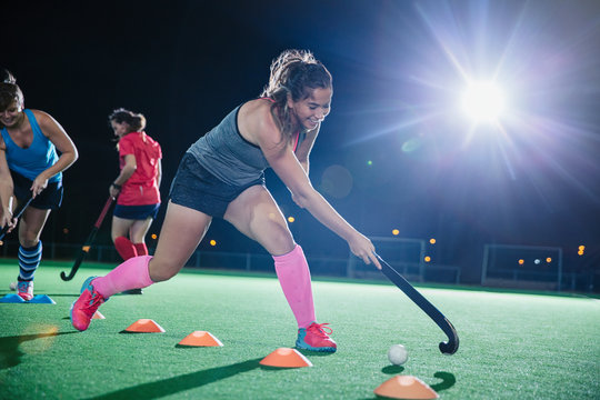 Determined young female field hockey player practicing sports drill on field at night