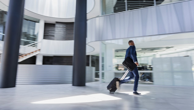 Businessman rushing, pulling suitcase in modern office lobby