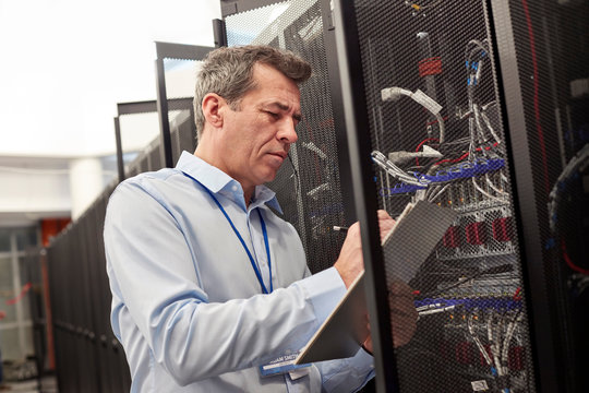 Male IT technician writing on clipboard at panel in server room