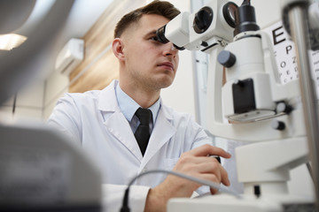 Low angle portrait of male optometrist using refractometer machine while testing vision of...