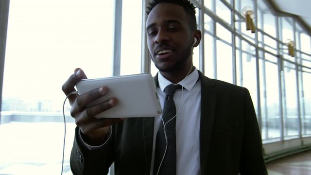 Pan of black businessman in suit and necktie wearing earphones and video chatting family on mobile phone while standing in hallway of office building with Panoramic windows