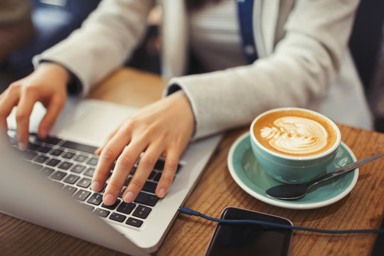 Hands of young woman using laptop, drinking cappuccino in cafe
