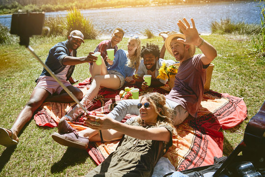 Playful young friends taking selfie selfie stick, enjoying picnic at sunny summer riverside