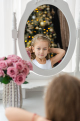 A little girl sits near a mirror and does a hairstyle before Christmas.