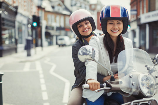 Smiling young women friends wearing helmets, riding motor scooter on urban street