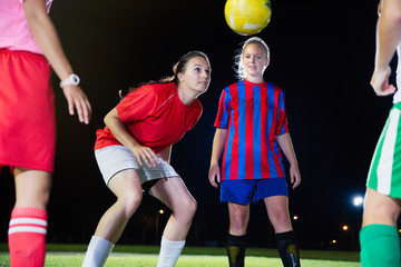 Young female soccer players practicing on field at night, heading the ball