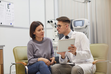 Portrait of young ophthalmologist using digital tablet while consulting female patient during eyesight check up in modern clinic, copy space