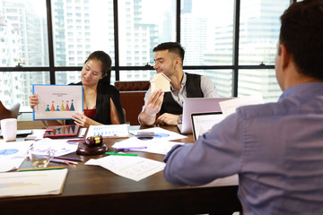 two caucasian businessmen having meeting with asian businesswoman. They use computer with chart, coffee cup and calculator beside while holding house model for new project with smiling face