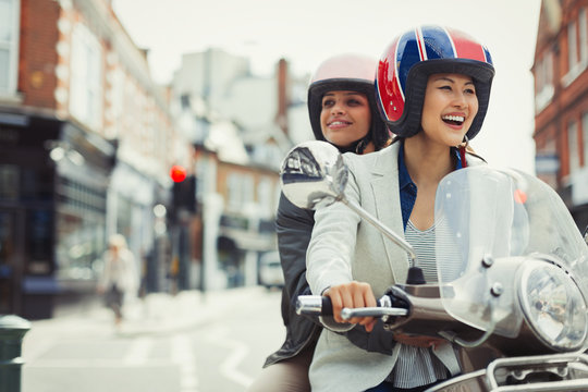 Smiling young women friends wearing helmets riding motor scooter on urban street