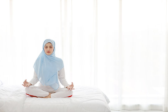 Young asian muslim woman sitting on bed and enjoying meditation. Beautiful girl in sleepwear with blue hijab practices yoga in bedroom with peace and calm. Healthy and lifestyle concept