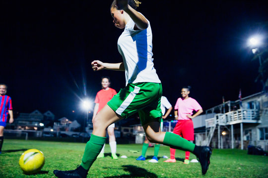 Young female soccer players playing soccer on field at night, kicking the ball