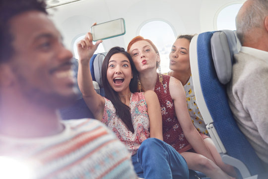 Playful young friends with camera phone taking selfie on airplane