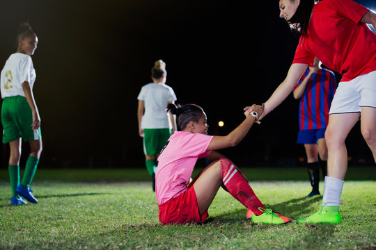 Fototapeta Young female soccer player helping fallen teammate get up on field at night