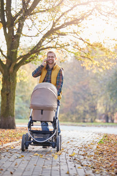 Smiling father talking on smart phone and pushing baby stroller in autumn park