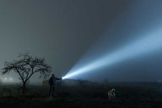 Man and dog exploring field at night, lightening with torch to sky at night.