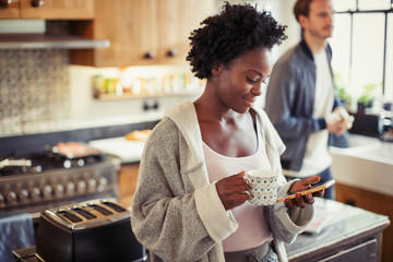 Woman drinking coffee, texting with smart phone in kitchen