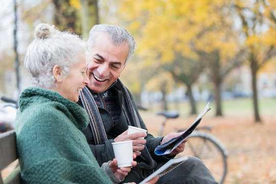 Smiling senior couple reading newspaper and drinking coffee on bench in autumn park