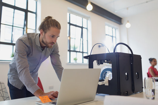 Male Designer Working At Laptop Next To 3D Printer In Office