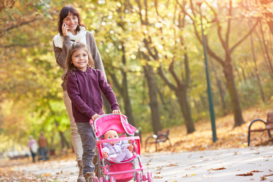 Smiling mother talking on smart phone with daughter pushing baby doll in stroller in autumn park