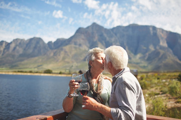 Romantic, active senior couple kissing drinking wine on sunny summer balcony at lakeside