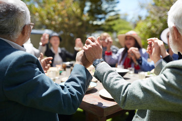 Active senior friends holding hands, praying at sunny garden party table