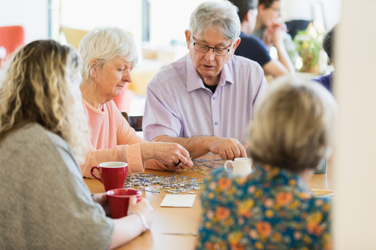 Senior friends assembling jigsaw puzzle drinking tea at table in community center - Powered by Adobe