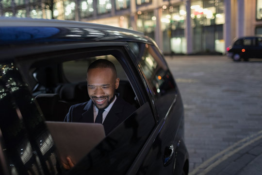 Smiling businessman using laptop in crowdsourced taxi at night