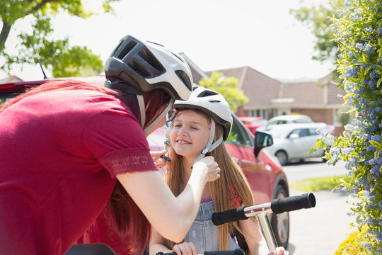 Mother fastening helmet on daughter riding scooter in sunny driveway