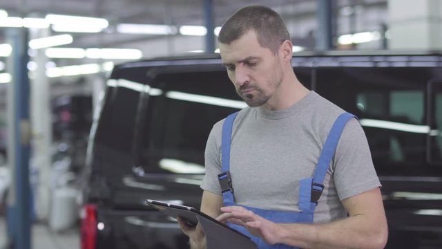 Young handsome Caucasian man in workwear standing with tablet at the background of black car. Serious male auto mechanic working in repair shop. Confidence, inspiration, lifestyle.