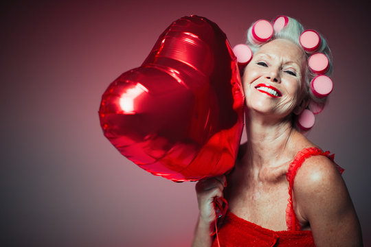 Portrait playful senior woman hair in curlers holding heart-shape balloon