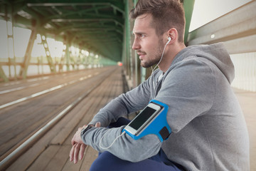 Young male runner resting, listening to music with headphones and mp3 player on train station platform