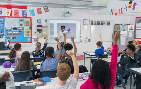 Male teacher leading lesson at projection screen in classroom students raising hands