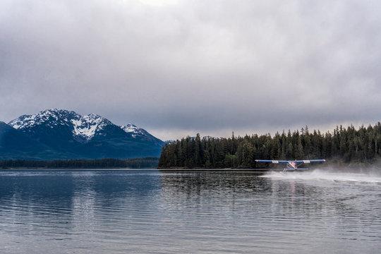 Float plane on the sea in Alaska. Snow peak mountains are on a background. Beautiful national park and green forest. Plane is getting in the air.