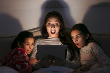 Surprised mother and daughters watching movie on digital tablet in dark bedroom