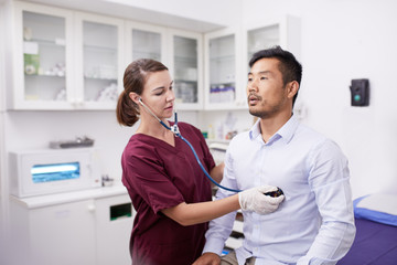 Female nurse using stethoscope on male patient in clinic examination room