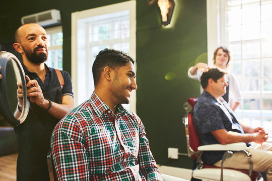 Male barber holding mirror for smiling customer in barbershop