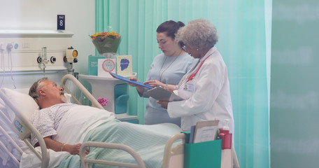 Doctors with medical chart making rounds, talking with senior patient in hospital room
