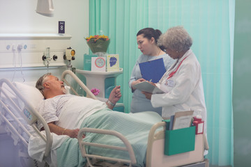 Doctors with medical chart making rounds, talking with senior patient in hospital room