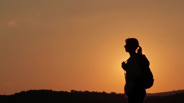Girl on the edge of the mountain as a silhouette in a beautiful sunset. Orange color. Silhouette of a girl with a backpack receiving sun dreds in the background. Medium plane. Epic shot