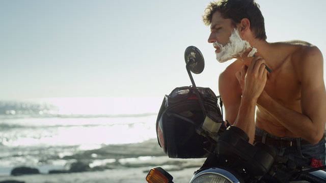 Young man on motorcycle shaving beard near ocean - Powered by Adobe