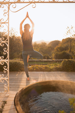 Woman practicing yoga tree pose on patio with hot tub and autumn tree view