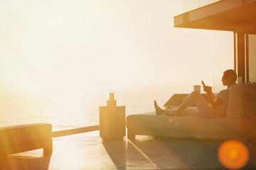 Silhouette woman using cell phone on chaise lounge on luxury balcony with sunset ocean view