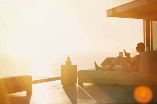 Silhouette woman using cell phone on chaise lounge on luxury balcony with sunset ocean view - Powered by Adobe