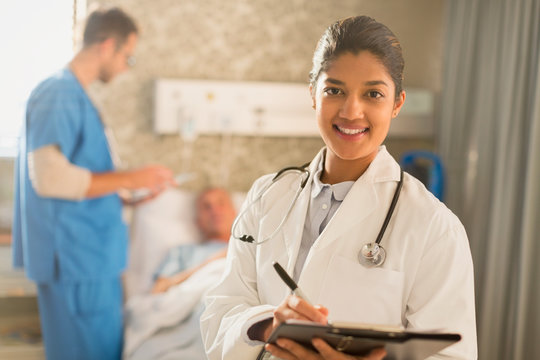 Portrait smiling female doctor making rounds, taking notes on medical record clipboard in hospital room